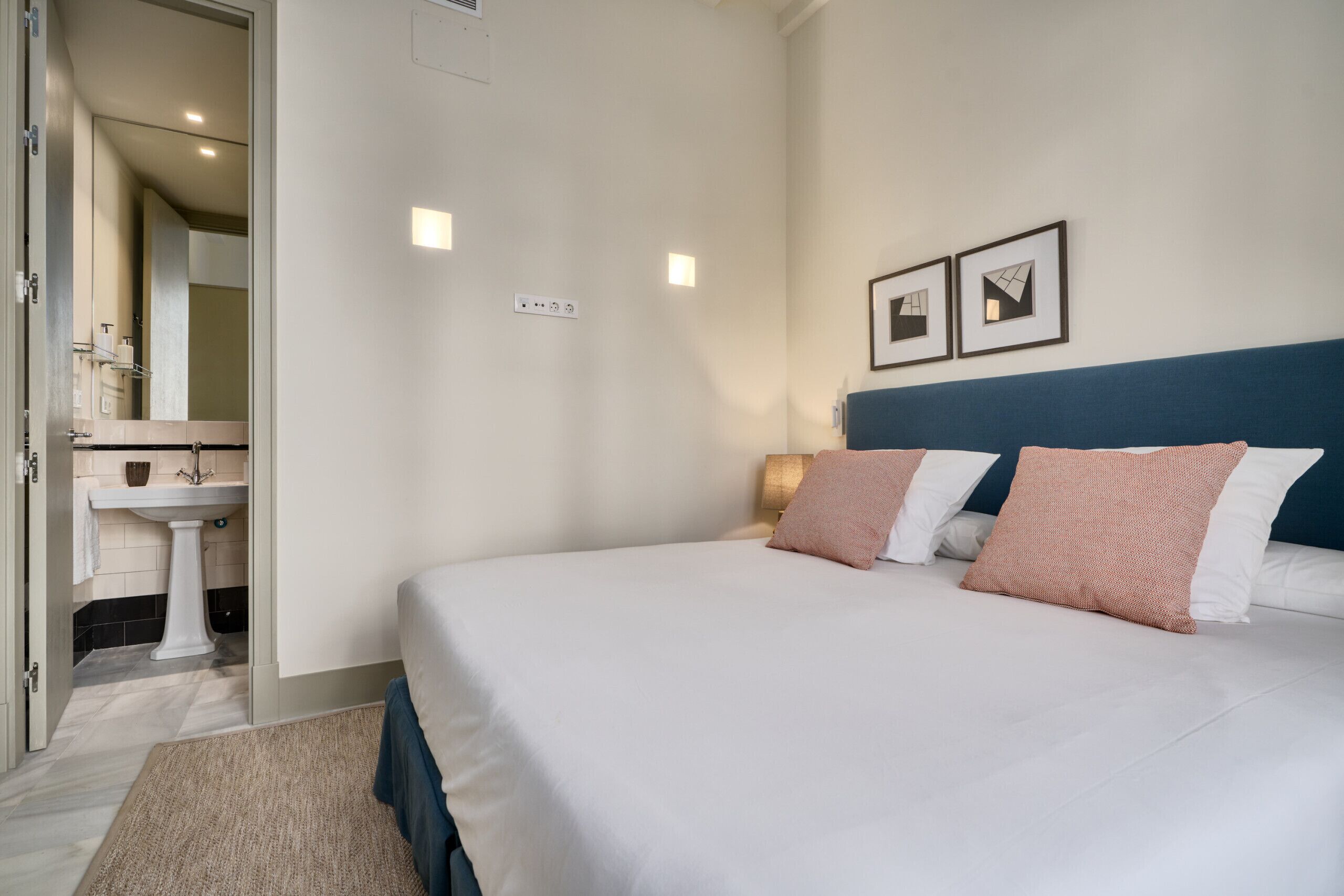 A bedroom with a bed, a bedside table, a nightstand, a wall-mounted light, and a door leading to a bathroom.