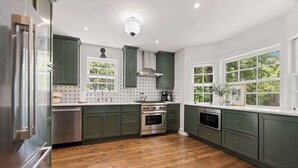 A kitchen with green cabinets, a stainless steel refrigerator, a built-in oven, and a window with a view of trees. - Holly House-1mi to UofA-0.5mi to RazorbackGreenway (Fayetteville)
