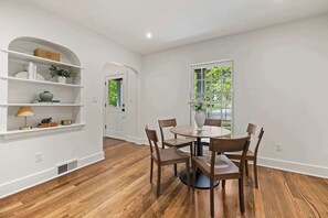 Un comedor con una mesa redonda y seis sillas, una pared blanca con un estante, y una ventana con vistas a follaje.