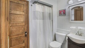 Kylpyhuoneessa on puinen ovi, valkoinen suihkuverho, valkoinen wc-istuin ja peilillä varustettu pesuallas.