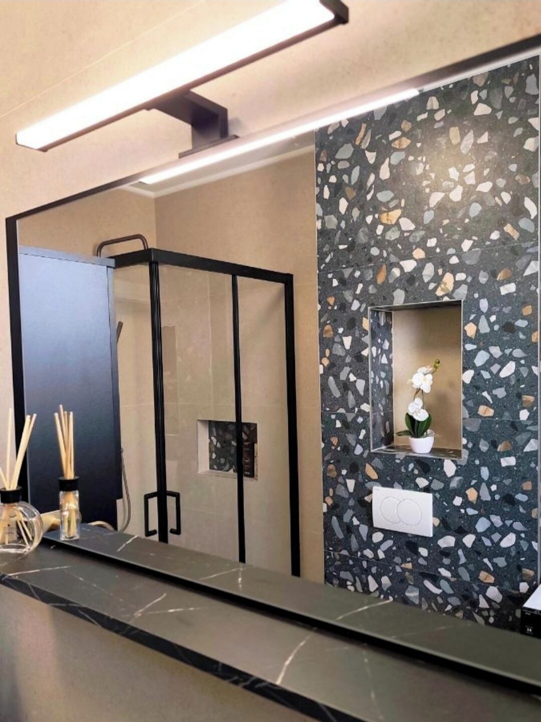 Et badeværelse med et spejl, et håndklædetørrestativ, en vask og en dekorativ væg med terrazzo-mønster.
