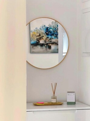 A wall-mounted circular mirror with a painting, a small shelf with a calendar, and a reed diffuser.