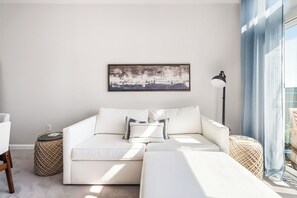 Una sala de estar moderna con un sofá blanco, una mesita auxiliar, una lámpara y un amplio ventanal con cortinas azules.