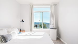 A bedroom with a large bed, a bedside table with a lamp, a window with curtains, and a wall-mounted air conditioner. - Elliott Bay View Condo Near Everything and Seattle Center with AC and More! (Seattle)