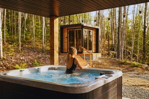 Eine Person in einem Whirlpool mit einer Holterrasse und einer Glaseinfriedung in bewaldeter Landschaft.