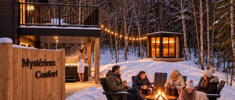 A cozy outdoor seating area with a fire pit, surrounded by snow, in front of a wooden building labeled 'Mystérieux Confort'.