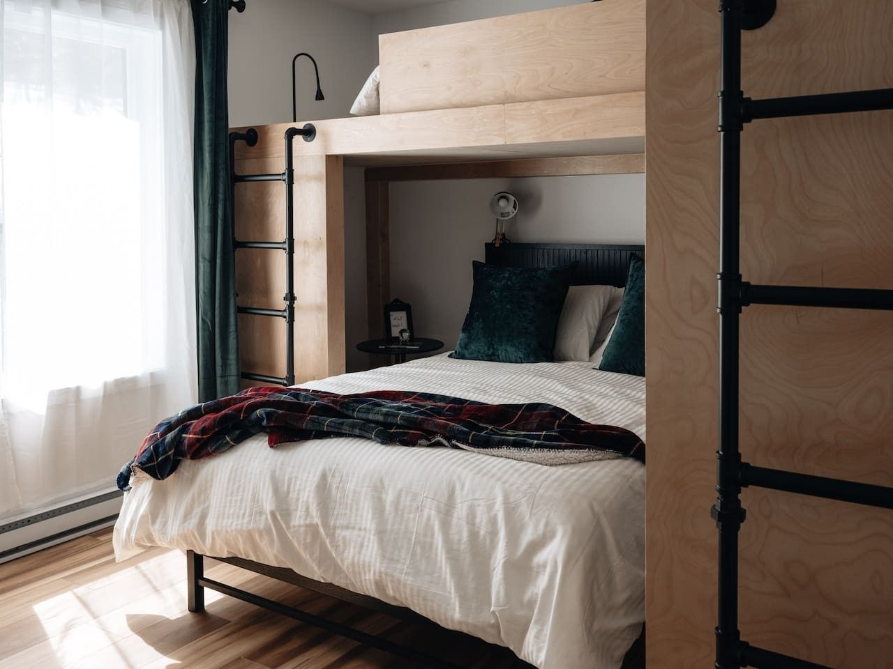 A bunk bed with a wooden headboard, a bedside table with a lamp, and a window with sheer curtains.