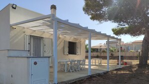 Un espacio de estar al aire libre cubierto, con una mesa y sillas, un pequeño armario de servicios y un edificio con contraventanas.