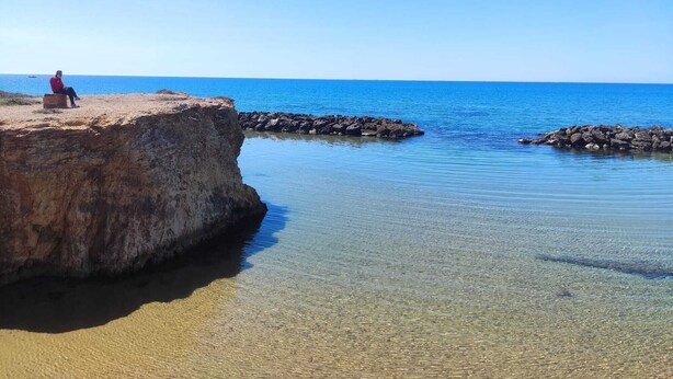 Escena costera con una persona sentada sobre una roca, agua azul cristalina y un promontorio rocoso.