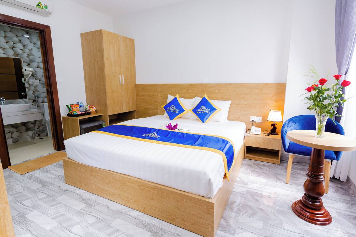 Ein Hotelzimmer mit Bett, Holzschrank, Nachttisch, Lampe, Sessel und einer Vase mit Blumen.