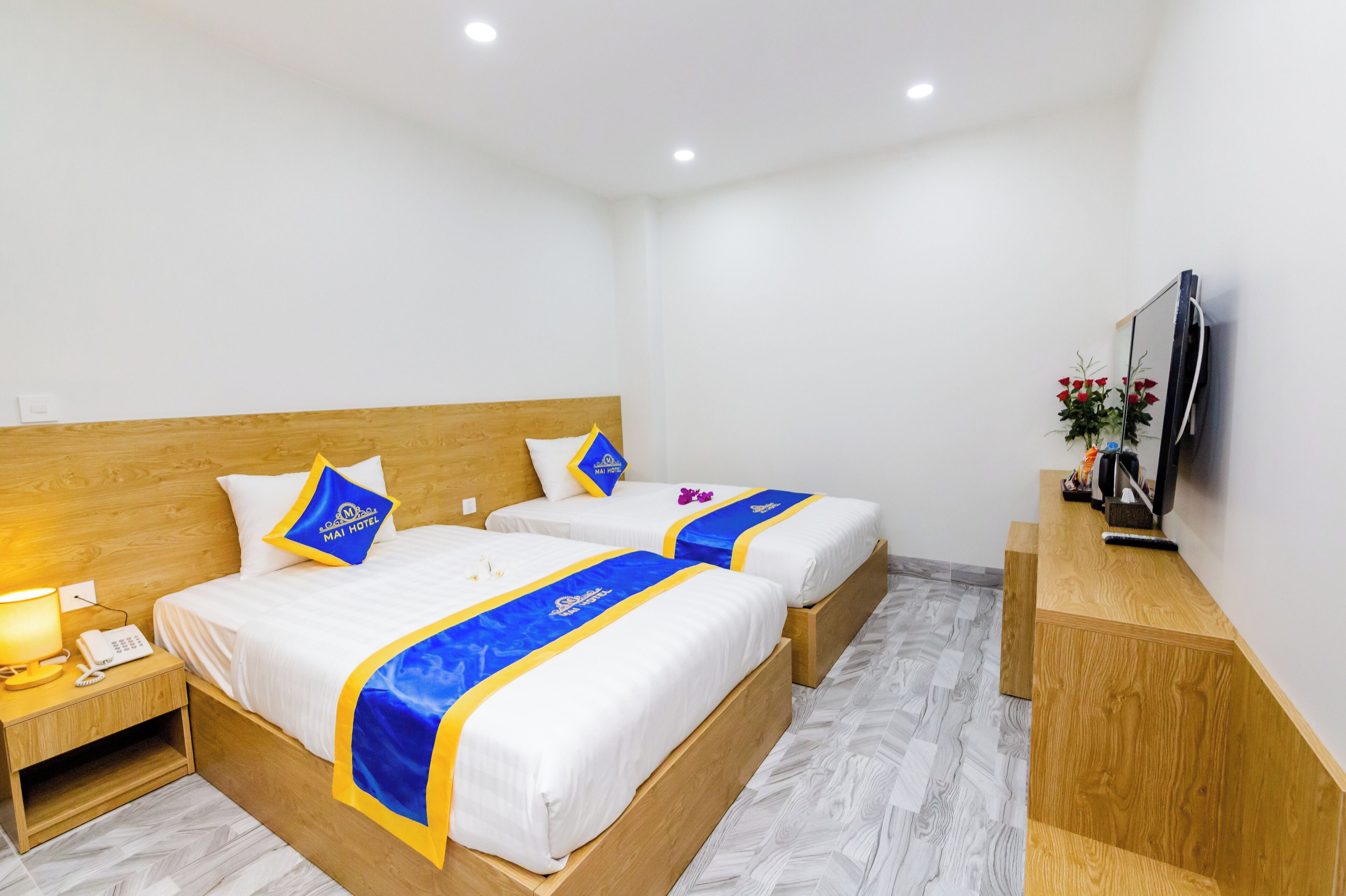 A hotel room with two beds, wooden headboards, a nightstand with a lamp, and a TV on the wall.