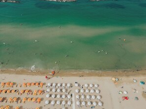 An aerial view of a beach with rows of umbrellas and sun loungers, a clear water body, and a rocky shoreline. - Mini House with Veranda (Mobile Home Camping) - Pools & Sea | Poseidon Resort (San Salvo Marina)