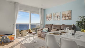 A modern living room with a view of the sea, a dining area with a table set for two, and artwork on the walls.