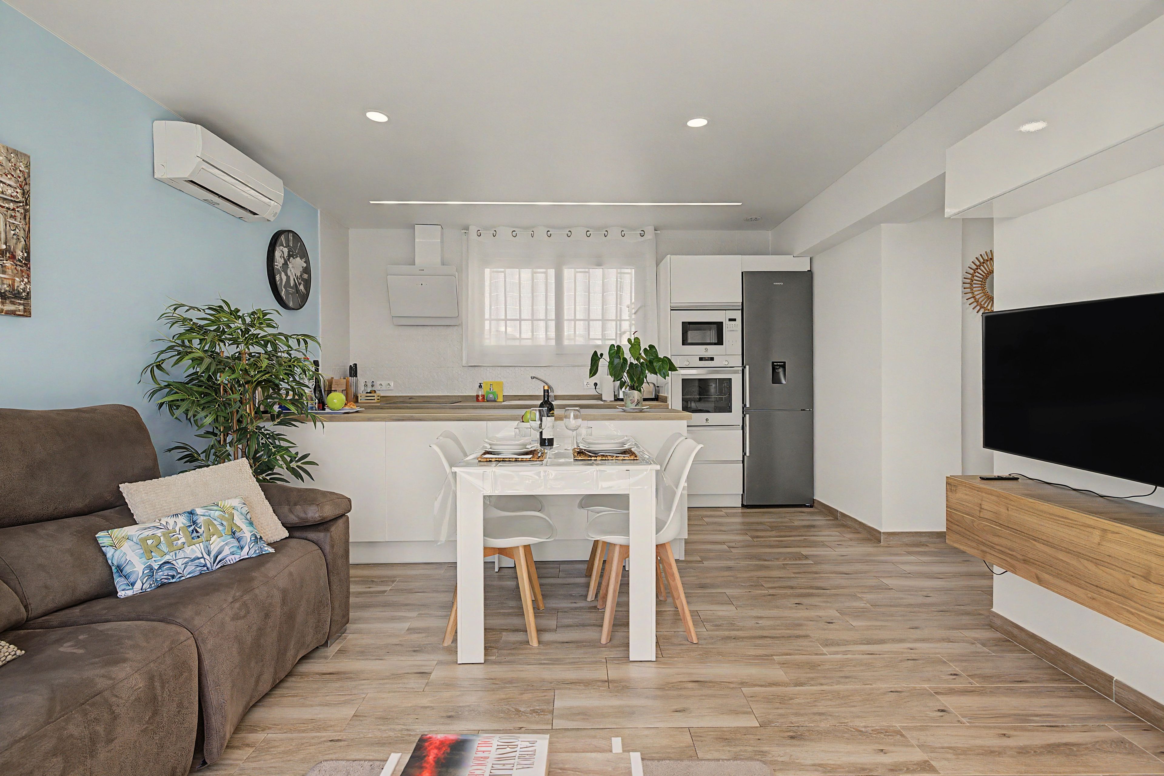 A modern living room with a brown sofa, a dining table with chairs, a flat-screen TV, and a kitchen area with white cabinets and appliances.