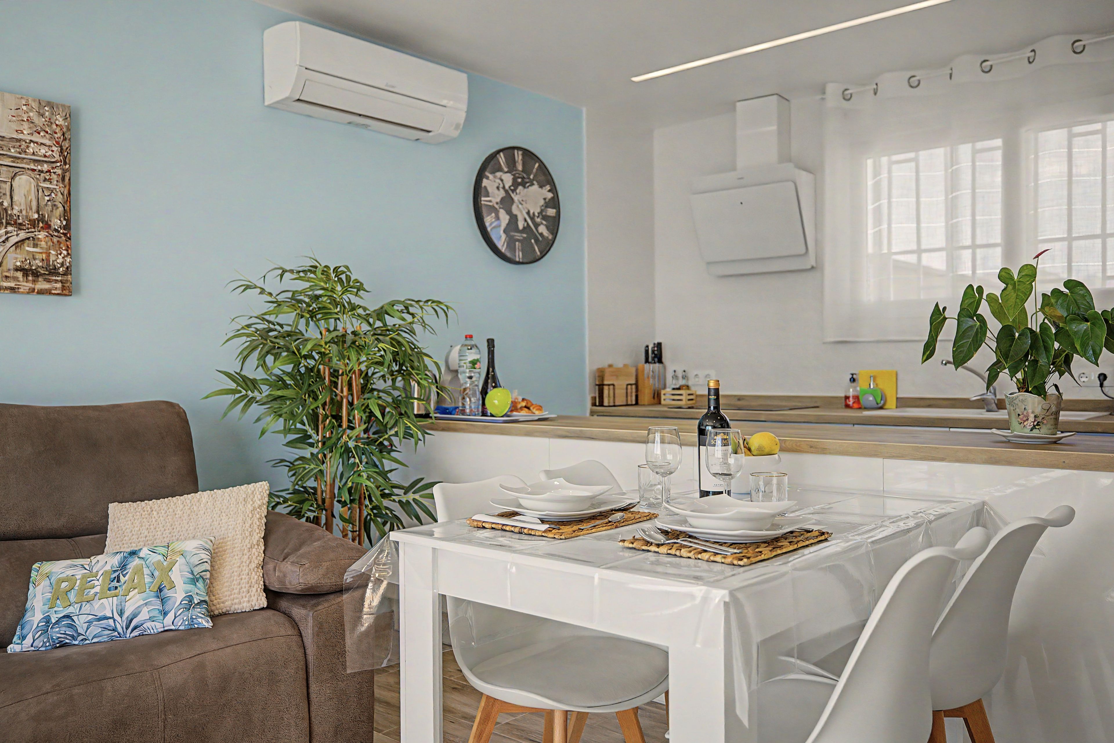 Un coin repas avec une table blanche dressée pour deux personnes, un canapé marron agrémenté d’un coussin où est inscrit le mot « RELAX », une grande plante verte et un climatiseur fixé au mur.