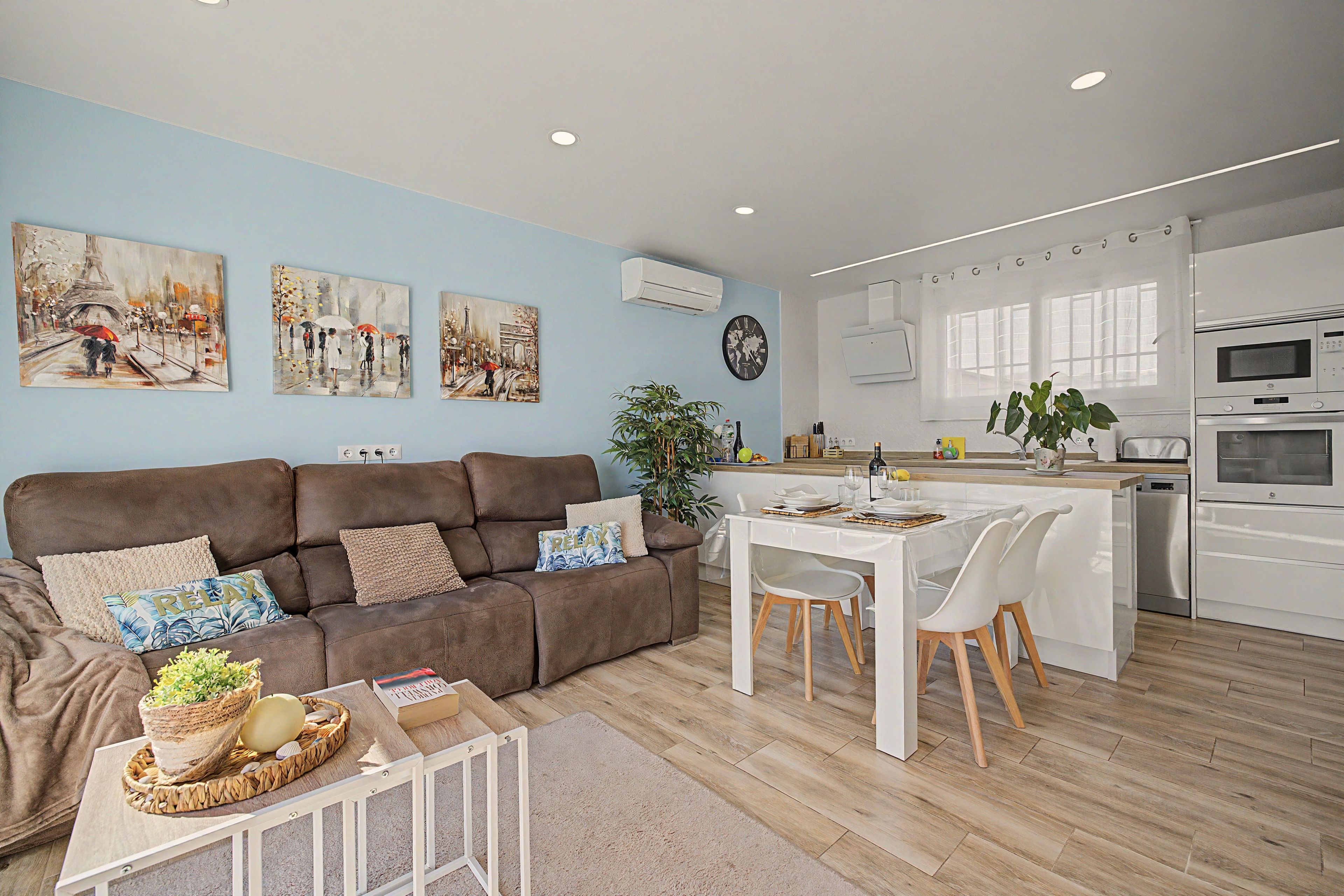 Een moderne woonkamer met een bruine bank, een eettafel met stoelen en een keuken met witte kasten en apparatuur.