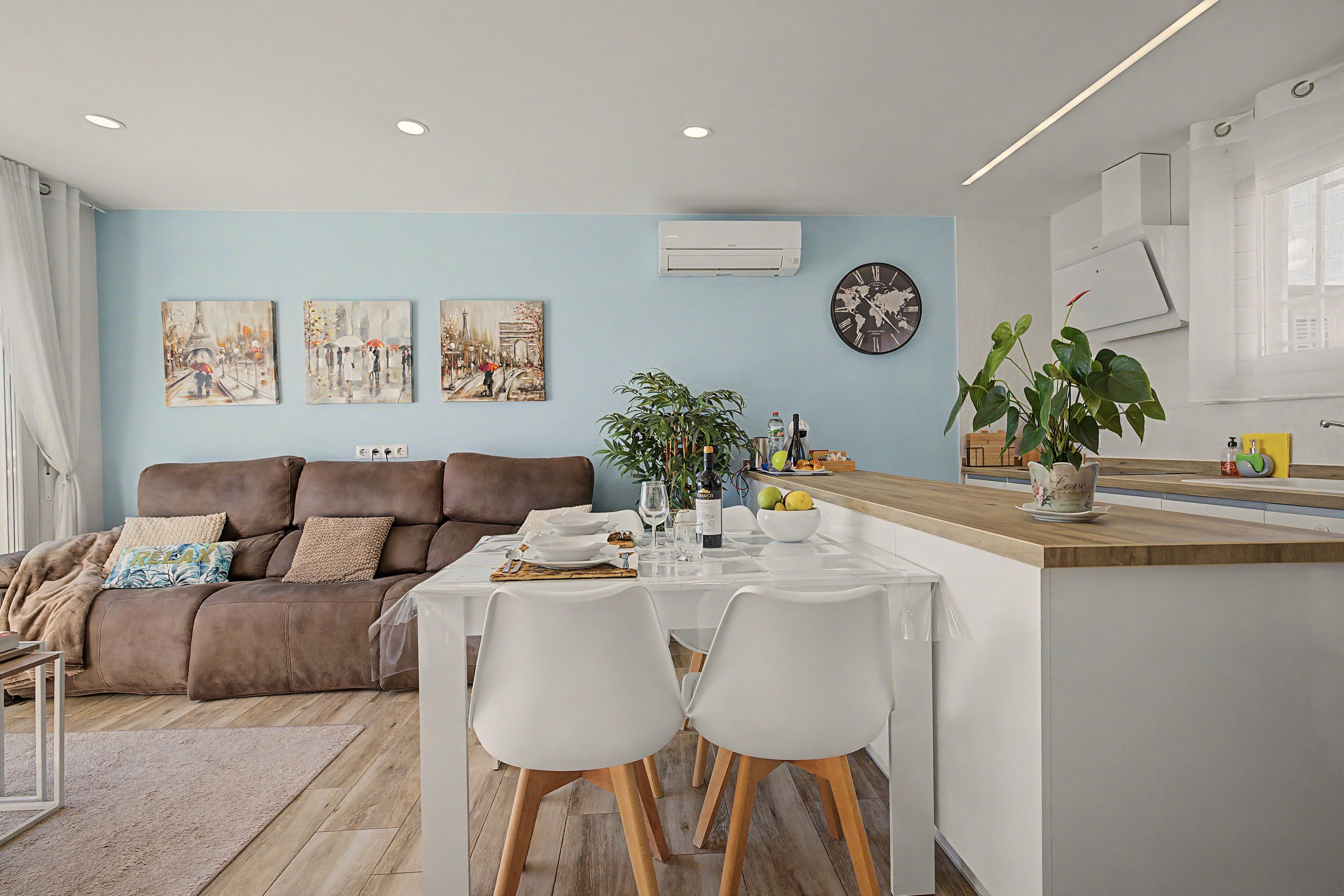 Un salon moderne avec un canapé marron, une table à manger dressée pour deux personnes et un coin cuisine doté d’un plan de travail en bois.