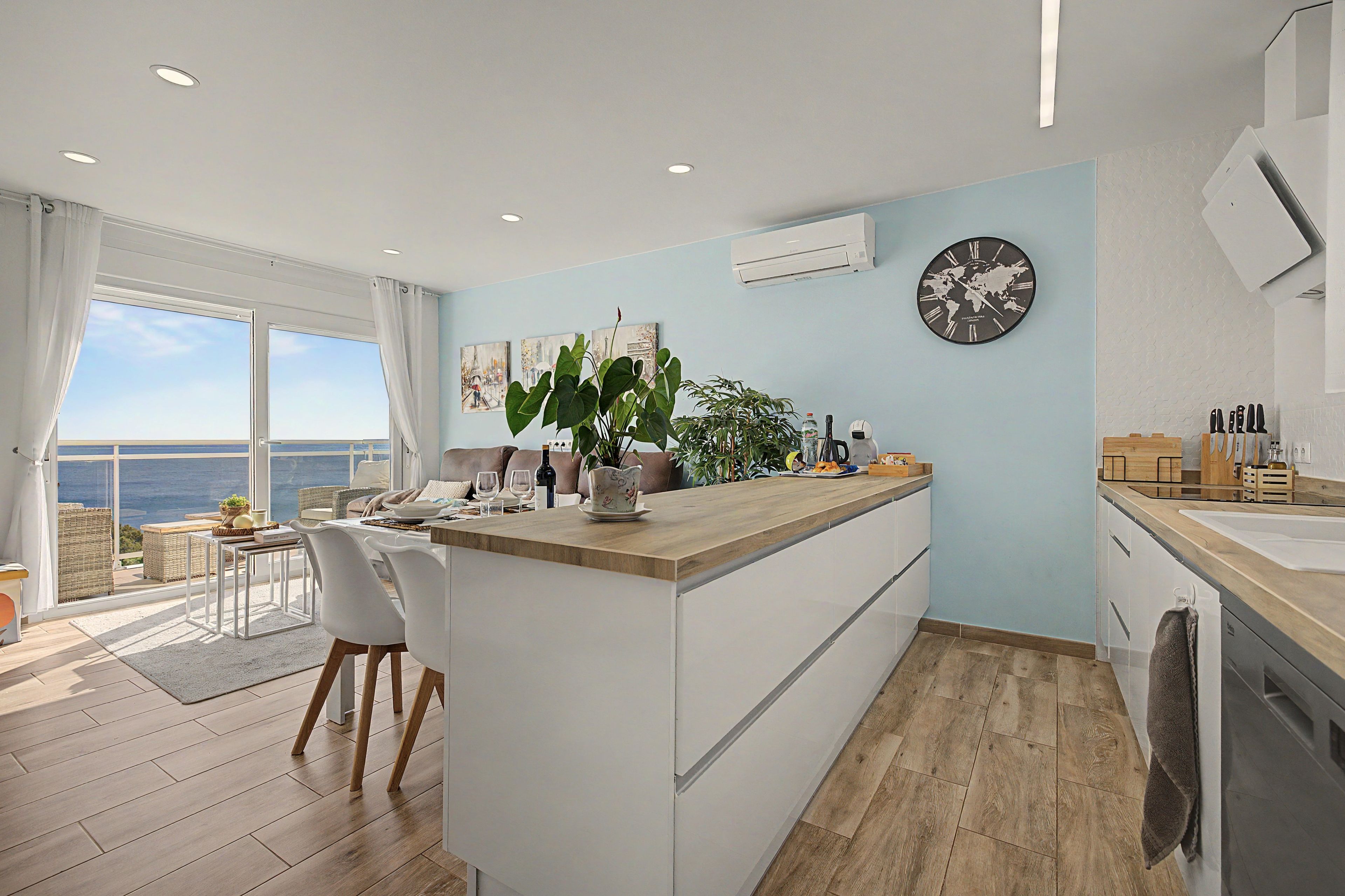 Een moderne keuken met een houten aanrecht, een eethoek met witte stoelen, en een groot raam met uitzicht op zee.