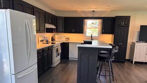 A modern kitchen with dark cabinetry, a white refrigerator, a microwave, a stove, and a bar area with stools. - Ingrid’s Green Haven - 27 Acre Private Farm (Wiarton)