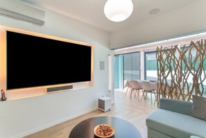 Un salon moderne doté d’un grand téléviseur à écran plat, d’une table basse ronde sur laquelle se trouve un saladier rempli de fruits, d’un canapé et d’un coin repas avec des chaises et une table.