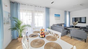 A modern living room with a dining area, a sofa set, a flat-screen TV, and a large window with curtains.
