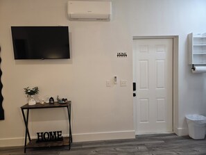 A room with a mounted TV, a white door with a 'HOME SWEET HOME' sign, a small table with decorative items, and a trash bin. - La cienfuegera suite 1 (Tampa)