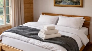 A wooden bed with white bedding, a mountain landscape painting above it, and a window with curtains. - Delightful 3-bedroom apartment in charming Frutigen awaits you (Frutigen)