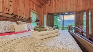 Quarto de cabana de madeira com cama, almofadas, cobertor, toalhas e vista pela janela.
