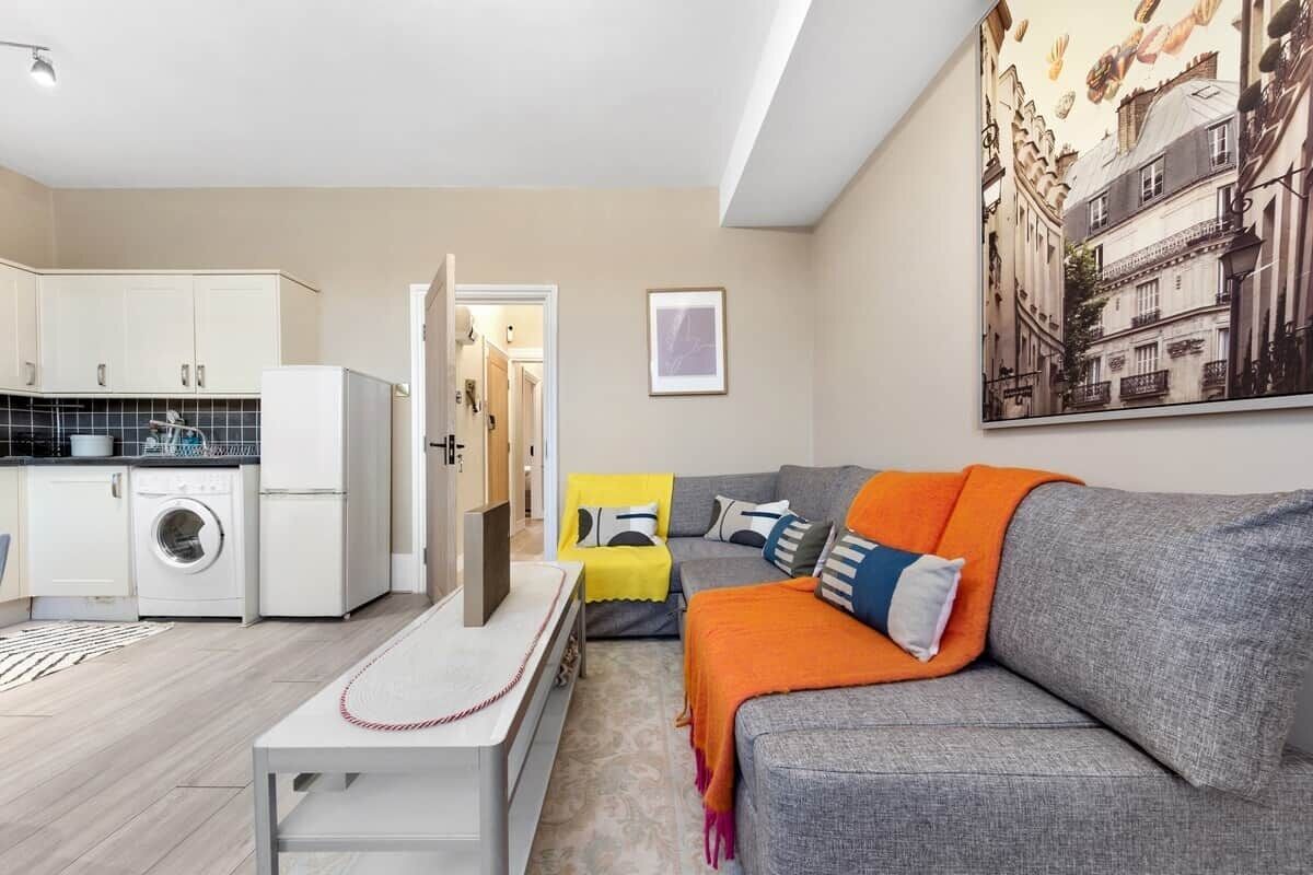 Um apartamento moderno com cozinha, um sofá cinza com manta laranja, um quadro emoldurado e uma obra de arte com vista para a cidade.