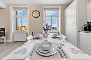 Una zona de dinar amb una taula preparada per a dues persones, un ram de flors i vista cap a edificis a través de les finestres.