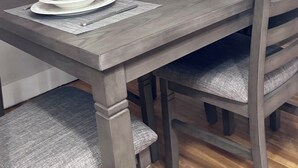 Una mesa de comedor con platos blancos y posavasos grises, rodeada de sillas de madera con cojines grises.