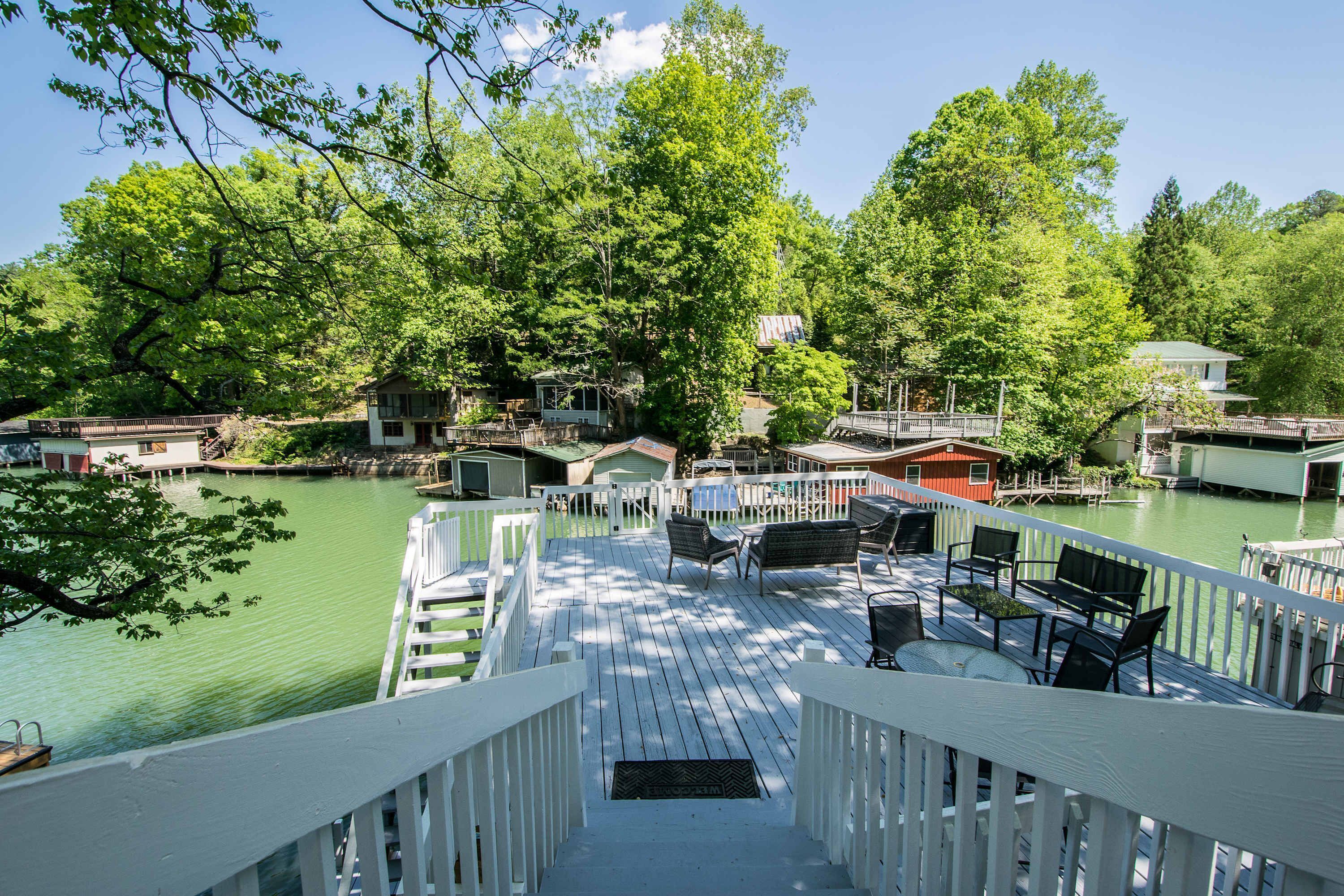 Une terrasse dotée de mobilier d’extérieur, offrant une vue sur un plan d’eau et entourée d’arbres et de maisons.