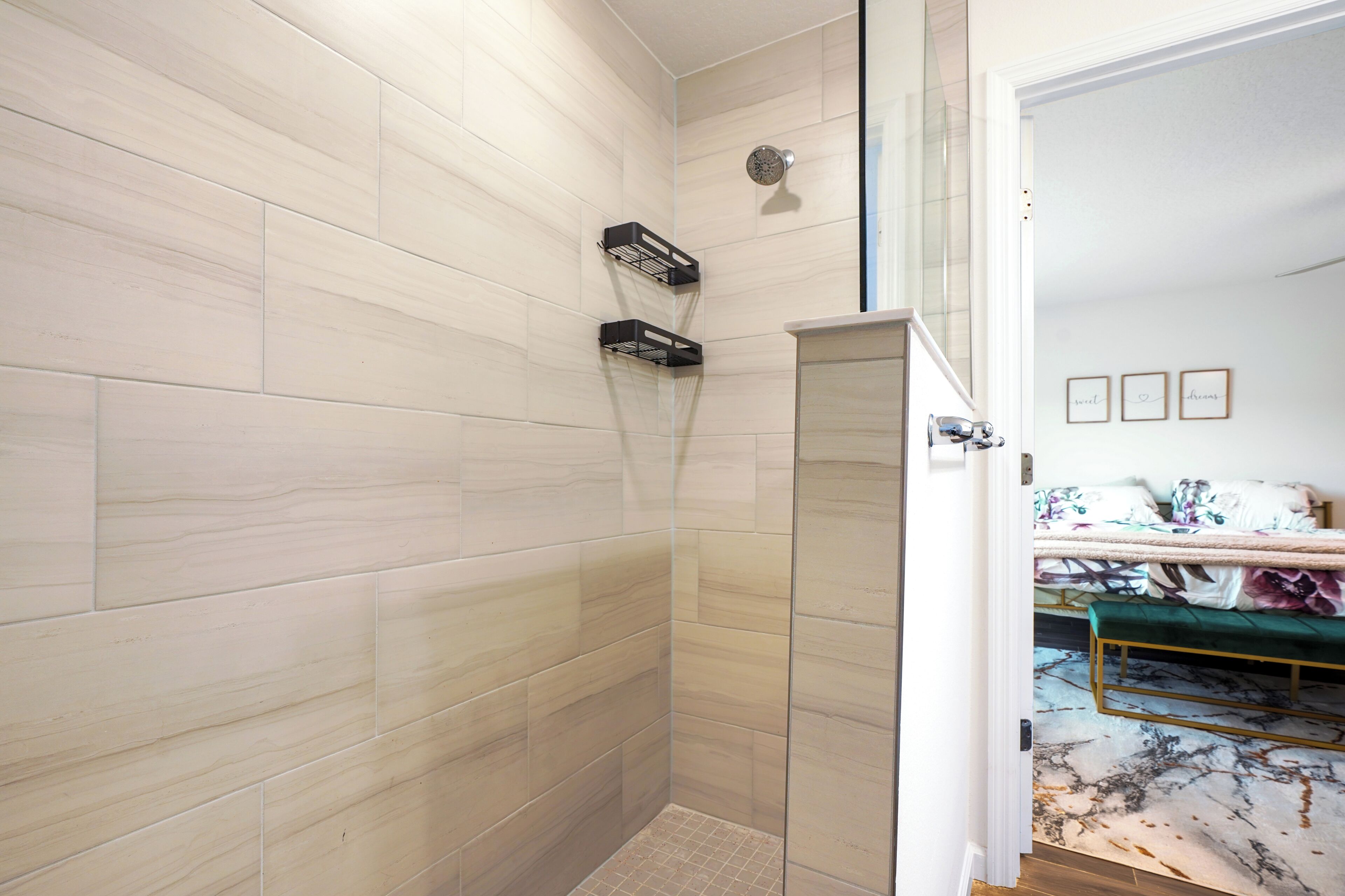 Een badkamer met een douche, een handdoekrek en een deur naar een slaapkamer met een bed en nachtkastje.