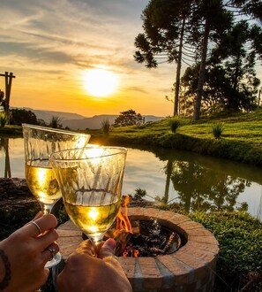 Zwei Gläser Weißwein in Händen, ein Feuerkorb und ein Sonnenuntergang über dem Wasser, Bäume im Hintergrund.