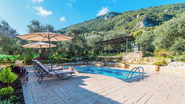 A pool with lounge chairs and umbrellas, surrounded by stone walls and greenery, with mountains in the background. - Villa Taxiarchis by Corfuescapes  (Corfu)