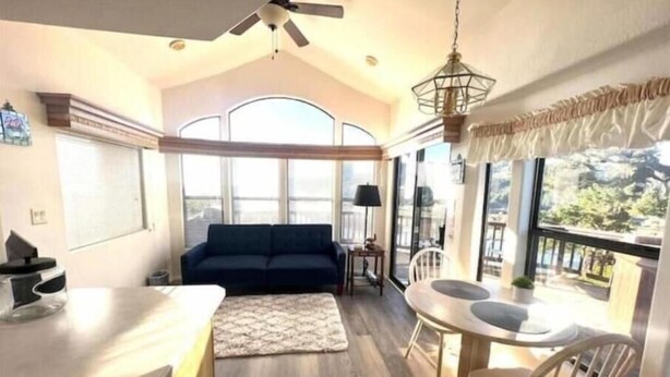 A spacious living area with a sofa, a dining table, and chairs, a ceiling fan, and large windows offering a view. - Rogue River Landing Resort (Gold Beach)