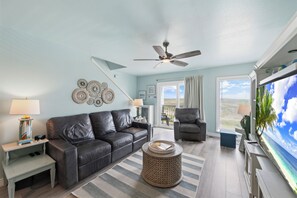 Ein Wohnzimmer mit Deckenventilator, einer Couch, einem Sessel, einem Fernseher und Blick auf den Ozean.