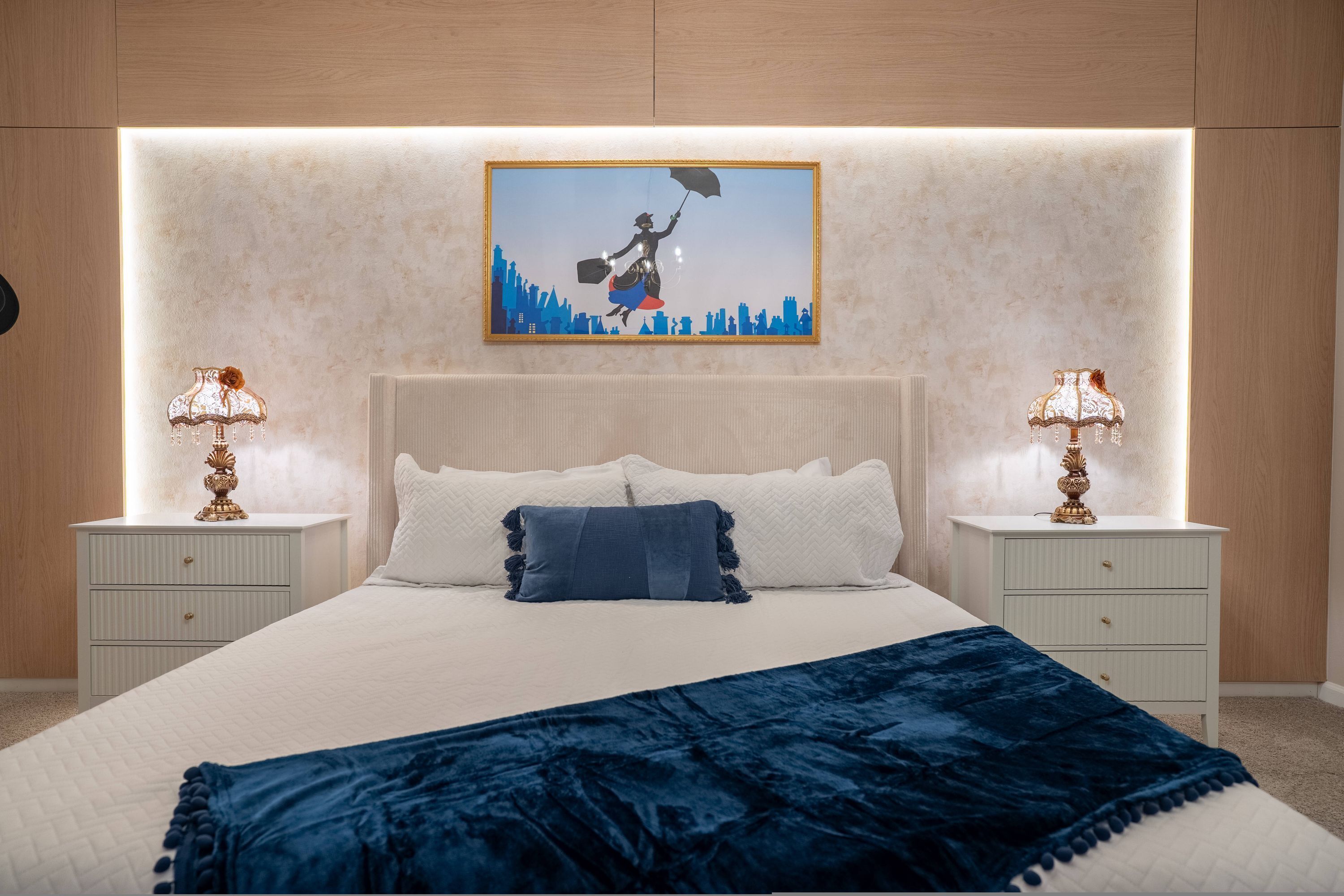 Un dormitorio con una cama grande, dos mesitas de noche con lámparas, un cuadro de una vista urbana en la pared y una manta azul.