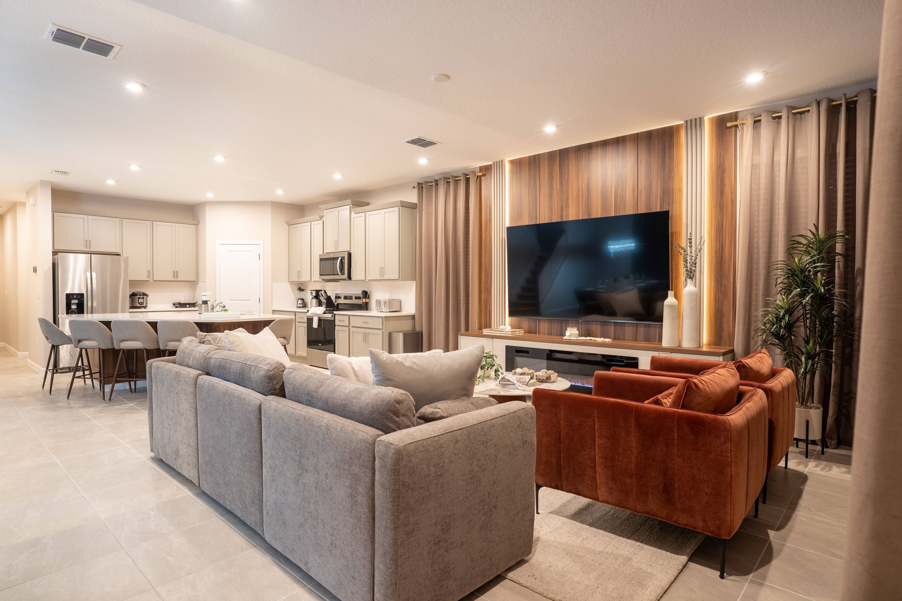 A modern living room with a large flat-screen TV, a grey sectional sofa, a red velvet sofa, and a kitchen in the background.