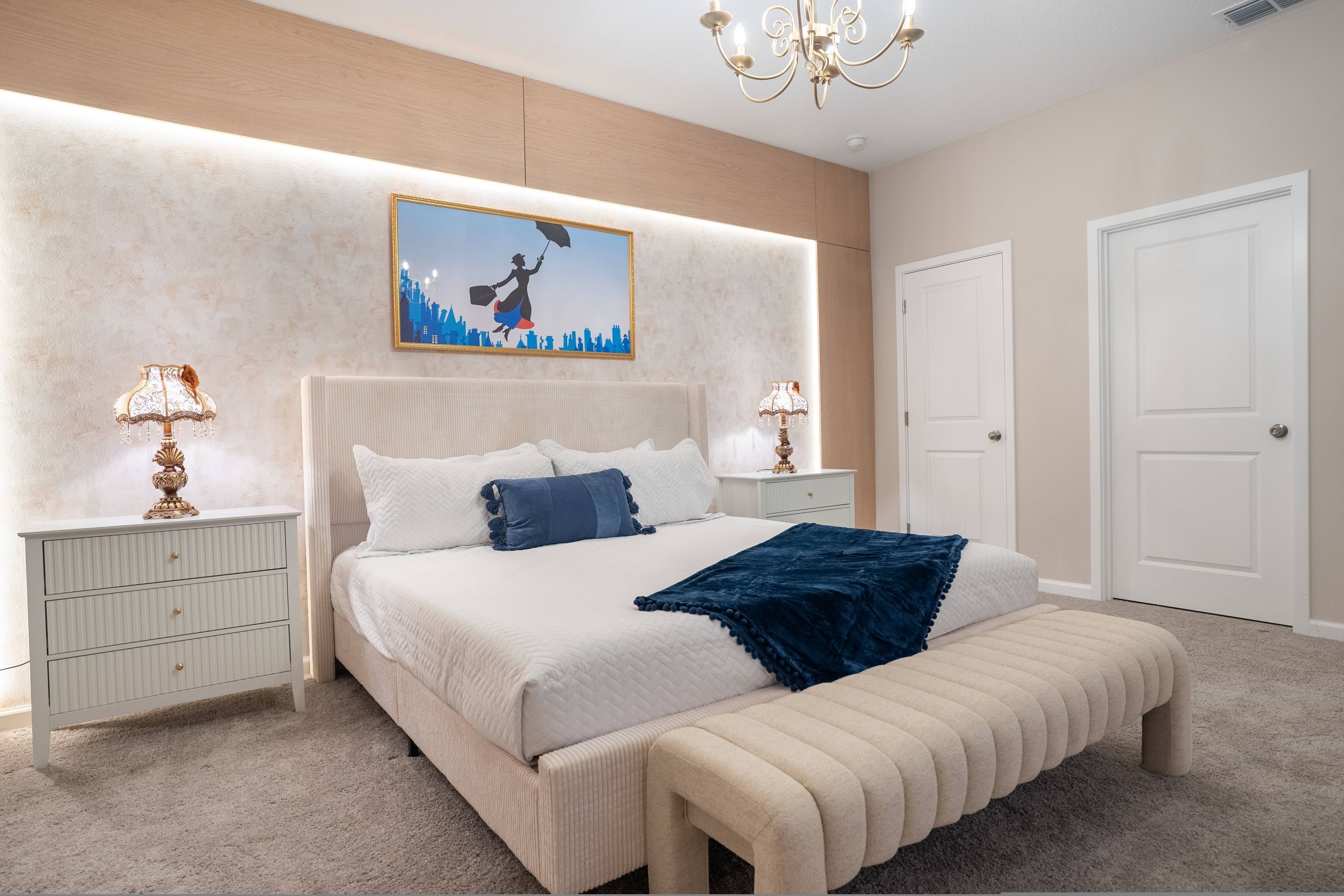 A bedroom with a large bed, two bedside tables, a chandelier, a painting, and a door.