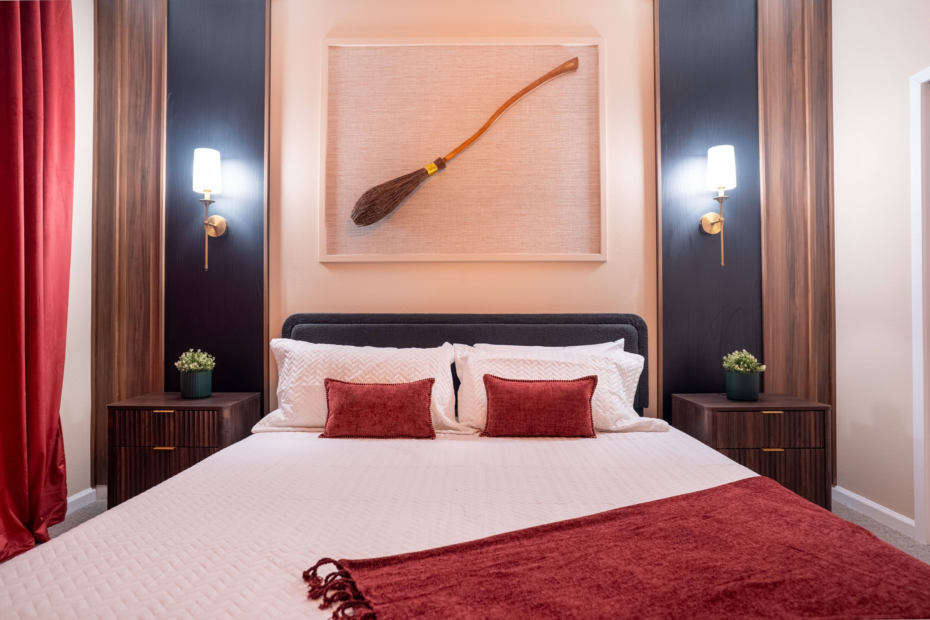 A neatly made bed with a red blanket, two bedside tables with lamps, and a wall-mounted picture of a broom.