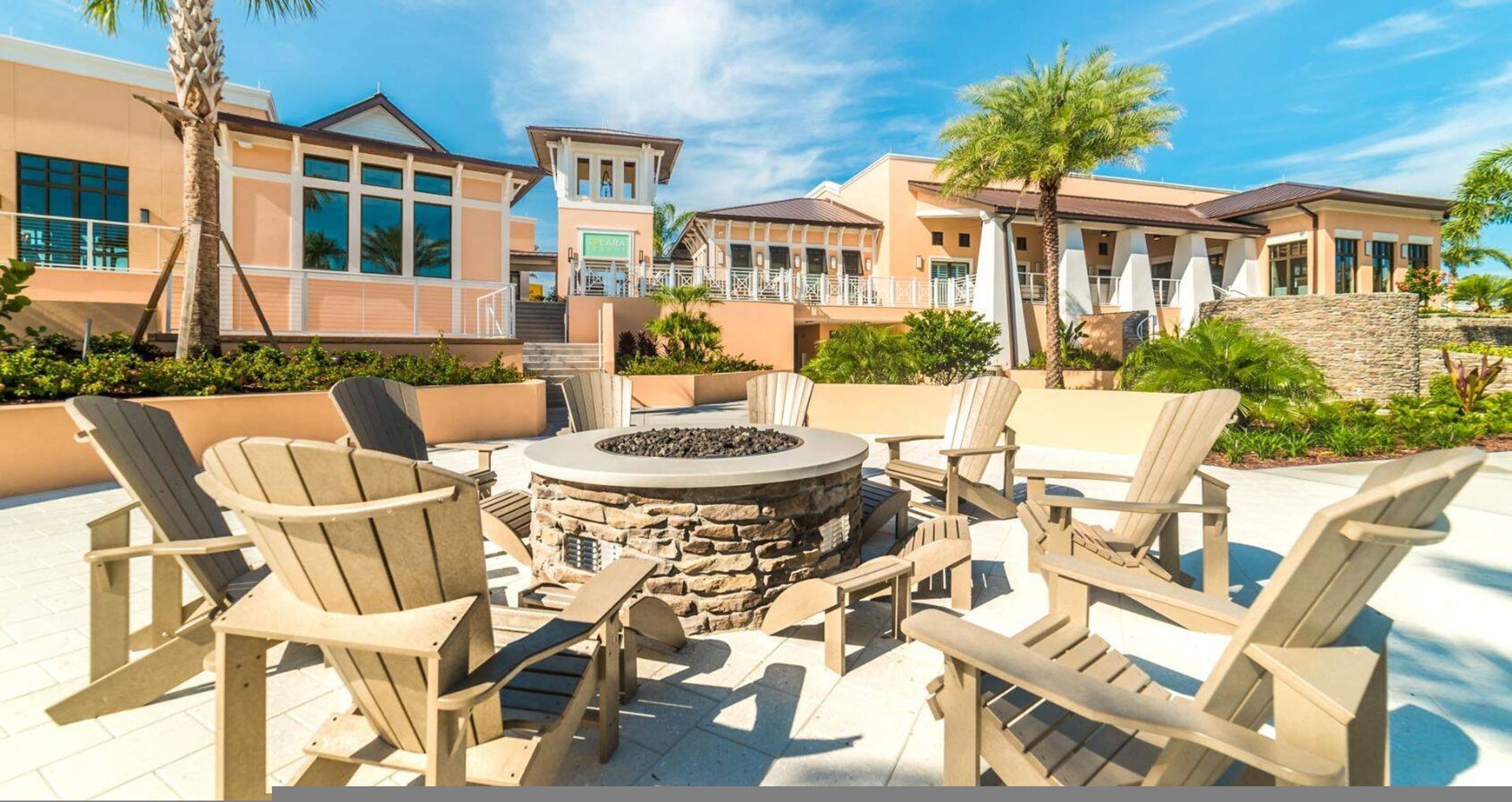 A patio area with Adirondack chairs, a stone fire pit, and a poolside view of a hotel with multiple buildings and a palm tree.