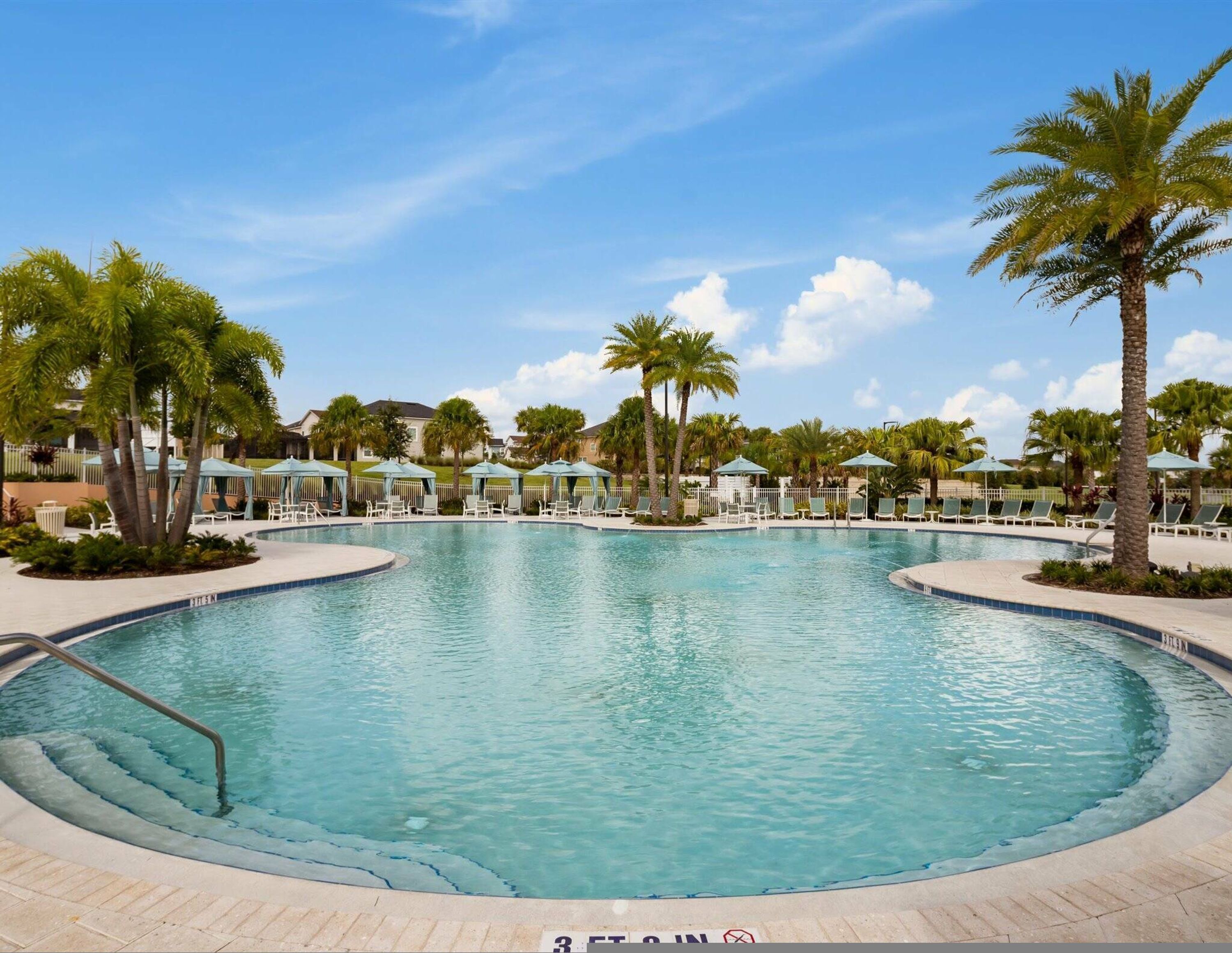 A large, clear swimming pool with lounge chairs and umbrellas, surrounded by palm trees and a clear sky.
