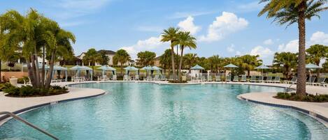 A large, clear swimming pool with lounge chairs and umbrellas, surrounded by palm trees and a clear sky.