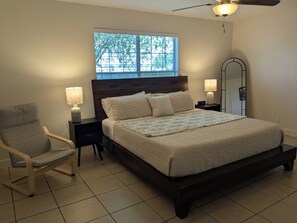 Un dormitorio con cama, sillón, mesita de noche, lámpara, ventana y ventilador de techo.