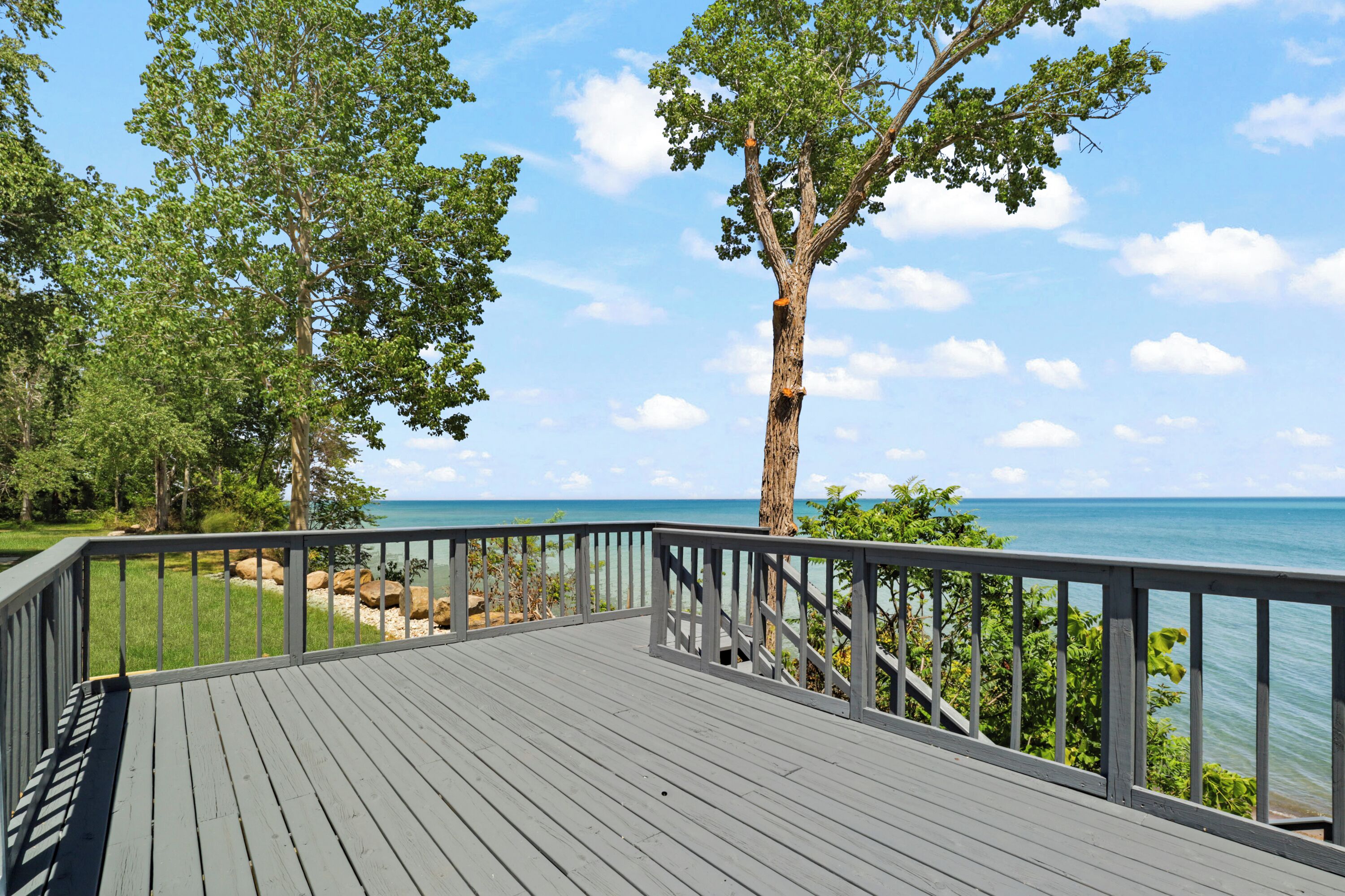 A wooden deck with a railing, overlooking a beach with clear blue water and a tree in the foreground.