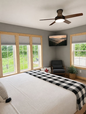 A bedroom with a bed, a chair, a ceiling fan, and large windows with a view of greenery.