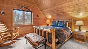 A log cabin bedroom with a bed, rocking chair, bench, and a window with a view of trees.