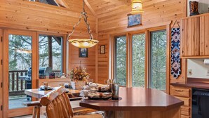 A wooden cabin interior with a dining area, kitchen, and large windows.