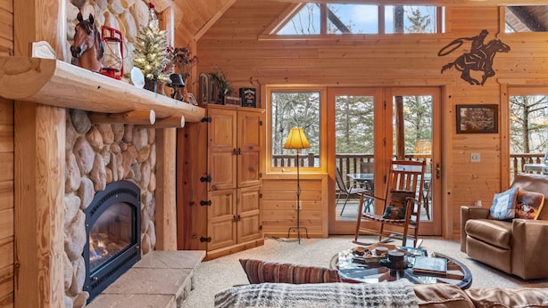 A cozy cabin interior with a stone fireplace, wooden shelves, a leather sofa, and a large window overlooking trees.
